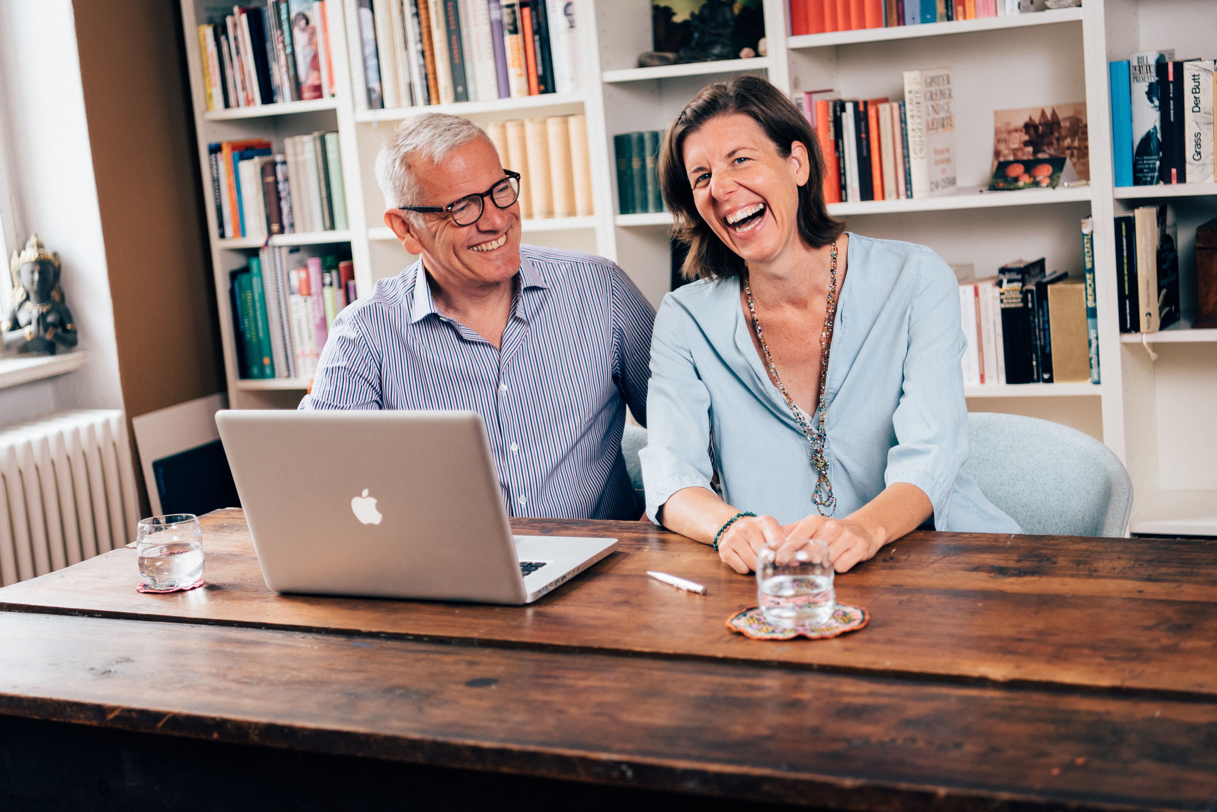 Trainer-Päarchen sitzt an einem Holztisch mit Laptop und Bücherwand und lacht in die Kamera